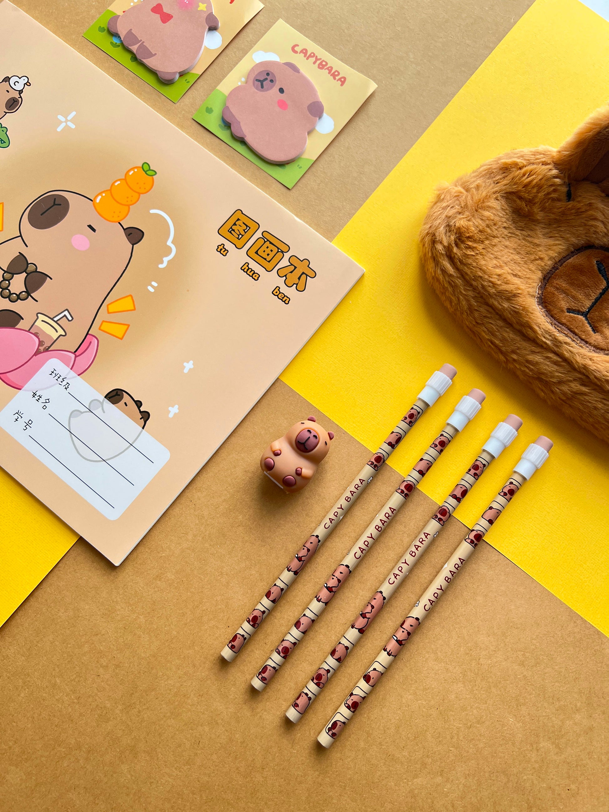 Close up of capybara themed pencils, sticky notes, and a fuzzy brown plush pencil pouch on a yellow background.